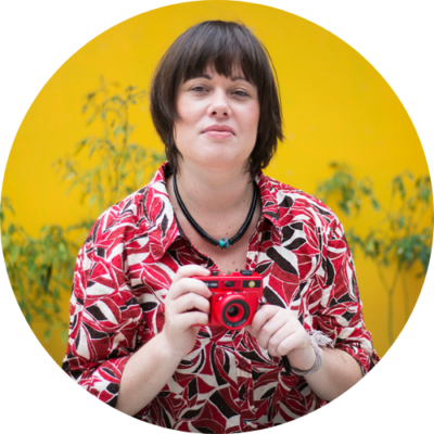 Willow Paule, content creator and strategist, wears red and holds a bright red Holga camera and stands against a bright yellow wall.