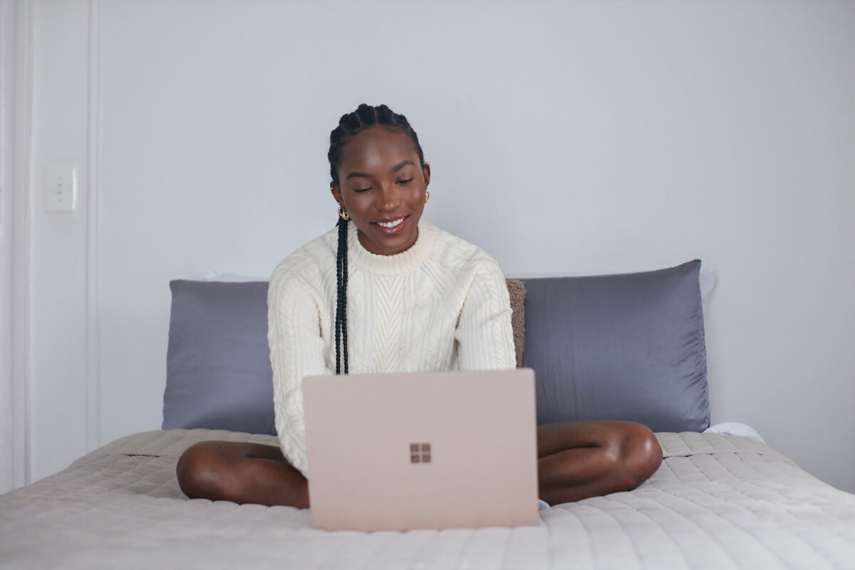 A Black women with braids who works from home, sits on her bed and smiles while choosing what collabs to pitch.