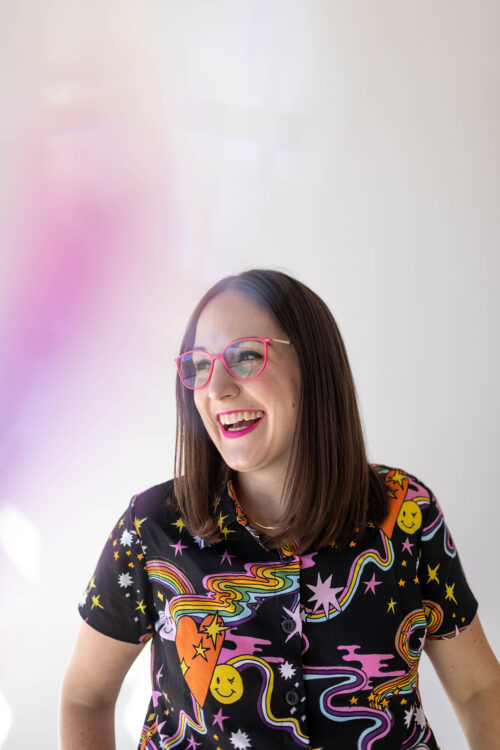 Kristy Black is wearing pink framed glasses, pink lipstick and an awesome rainbow and other shaped multicolor shirt.