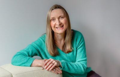 Sue Moore is a midlife white woman with blond hair; she wears a green sweater and smiles into the camera while leaning on the side of a beige couch.