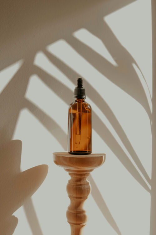 A brown bottle of skincare oil on a small wooden stand with dramatic plant shaped shadows on a beige wall behind it.