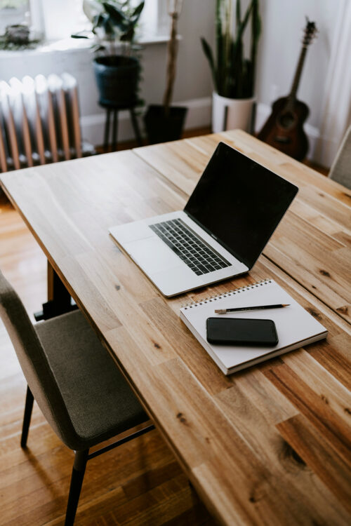 See this midlife woman's simple work setup: a wooden kitchen table, laptop, notebook, phone and pencil.
