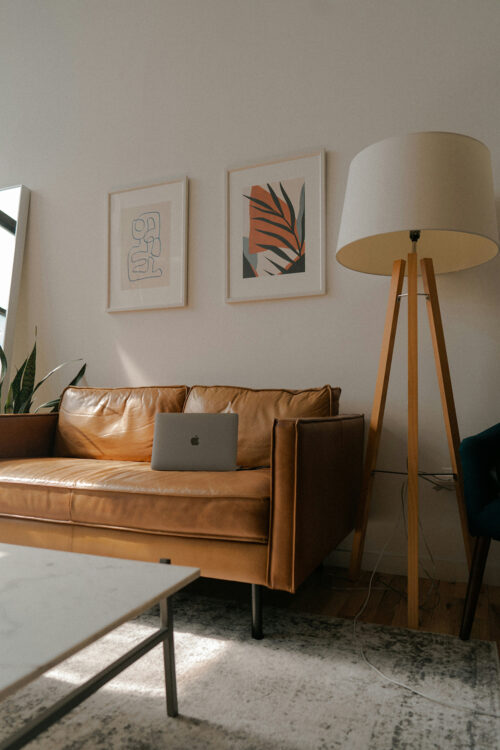 A moody, artsy living room with a pleather couch with a midlife woman's laptop open, a plant, and interesting illustrations on the wall.