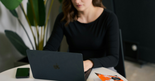 A woman in a black blouse checks her emails on a MacBook sitting at a table in front of a tropical green plant.